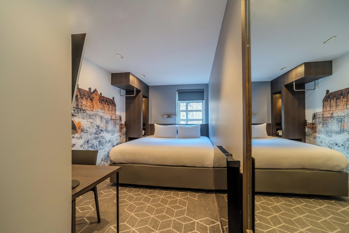 A modern hotel room with a large bed, a small table, and a window with a view of a building.