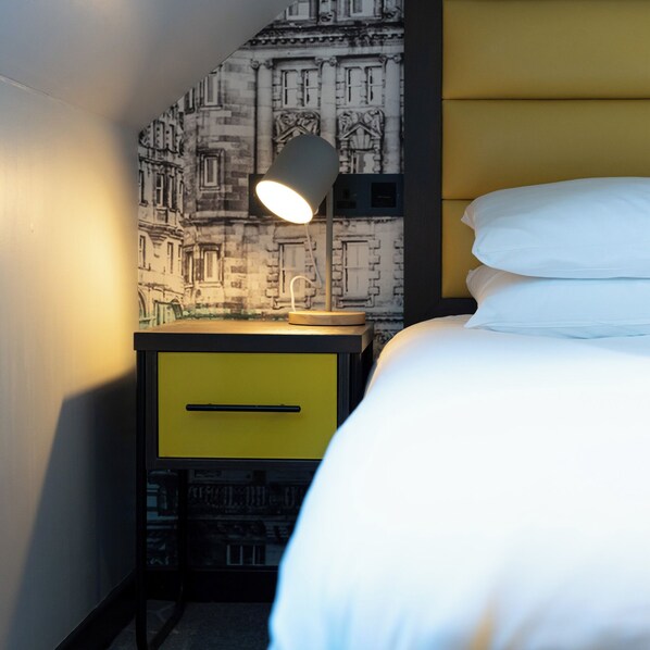 Quarto de hotel com cama, abajur, uma gaveta amarela e parede com mural de paisagem urbana.