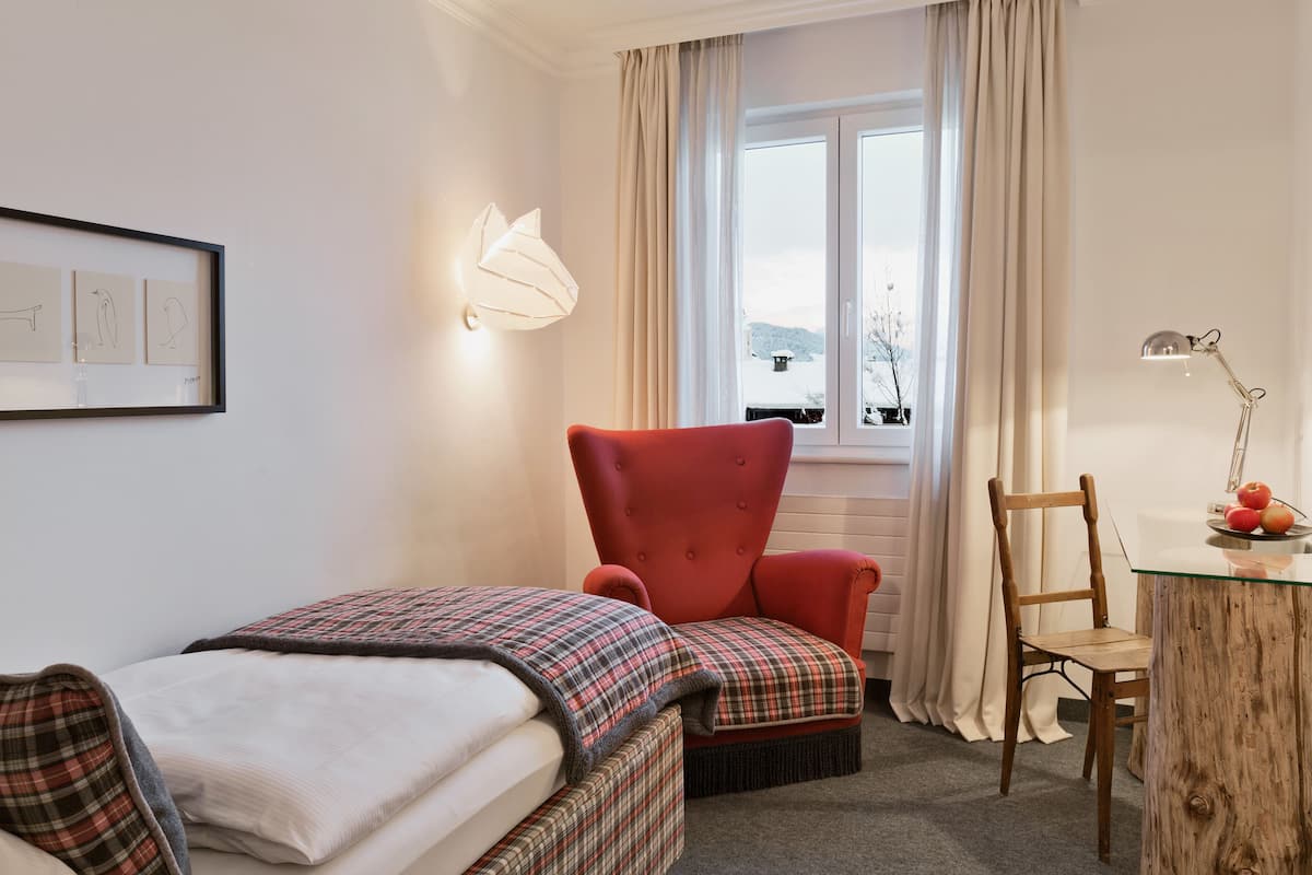 Una habitación de hotel acogedora con una cama, un sillón rojo, una silla de madera, una mesita de noche con una lámpara y una ventana con cortinas.