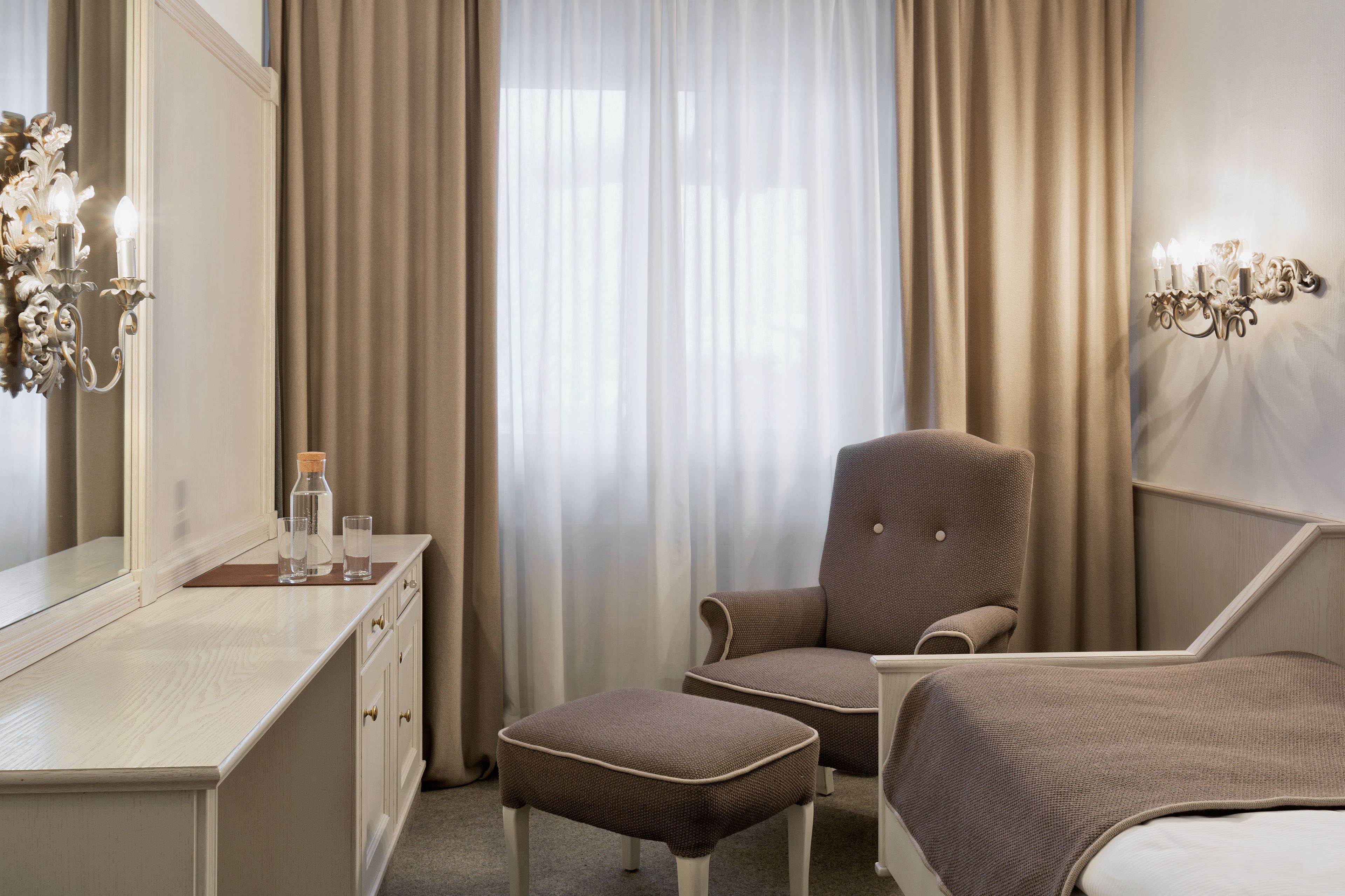 A hotel room with a bed, a chair, a desk with a mirror, and a wall-mounted light fixture.