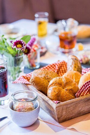 A table with a wooden tray holding several bread rolls, a small bowl of pickles, and a glass of red beverage. - Hotel Bruggwirt (Sankt Johann in Tirol)