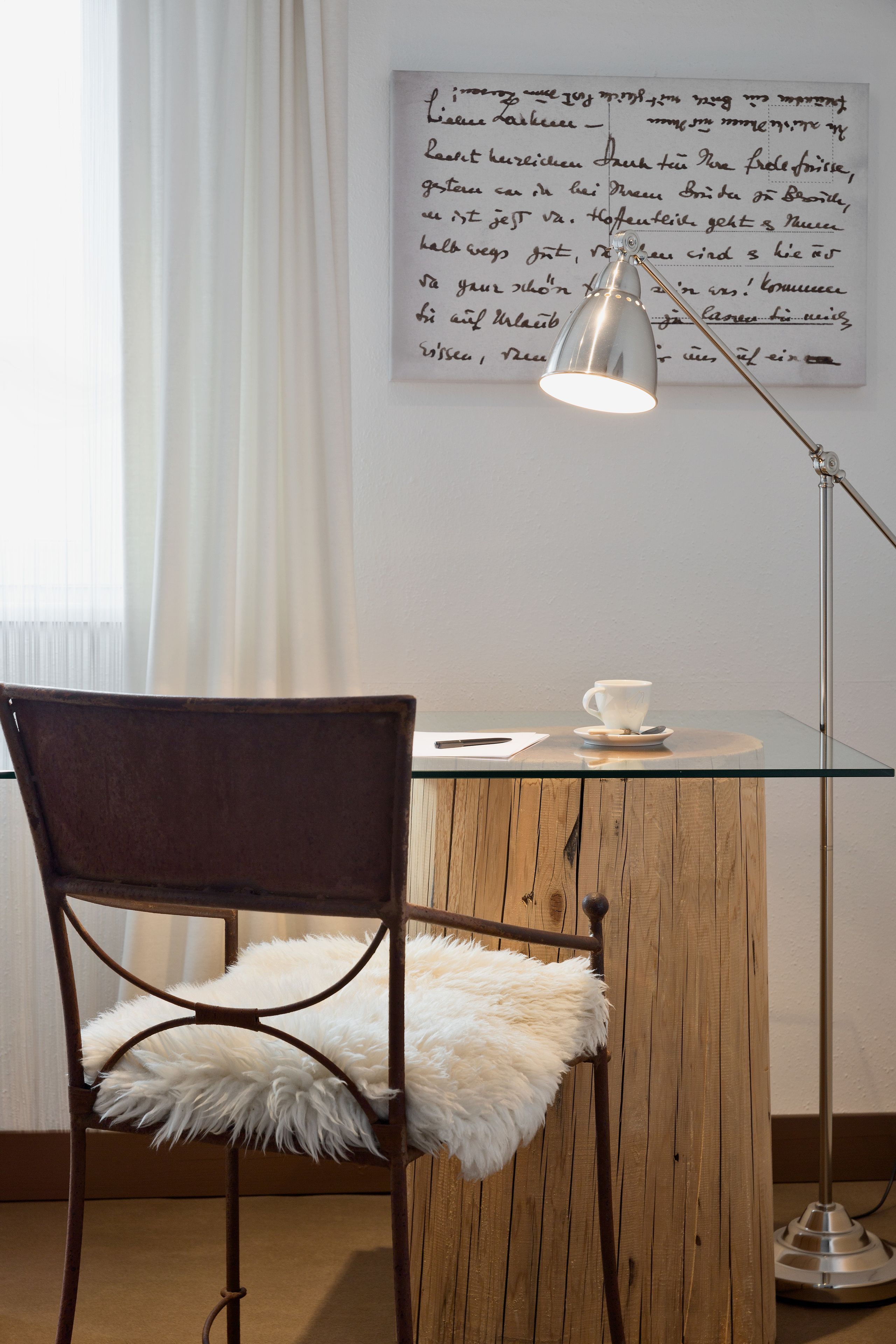 A chair with a fluffy white cushion, a glass table with a cup and saucer, and a floor lamp.