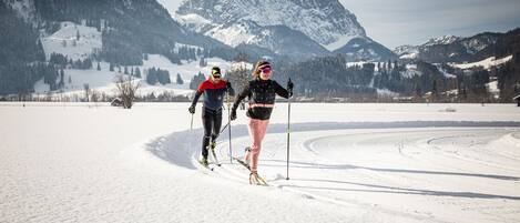 Two people cross-country skiing on a snowy field with mountains in the background.