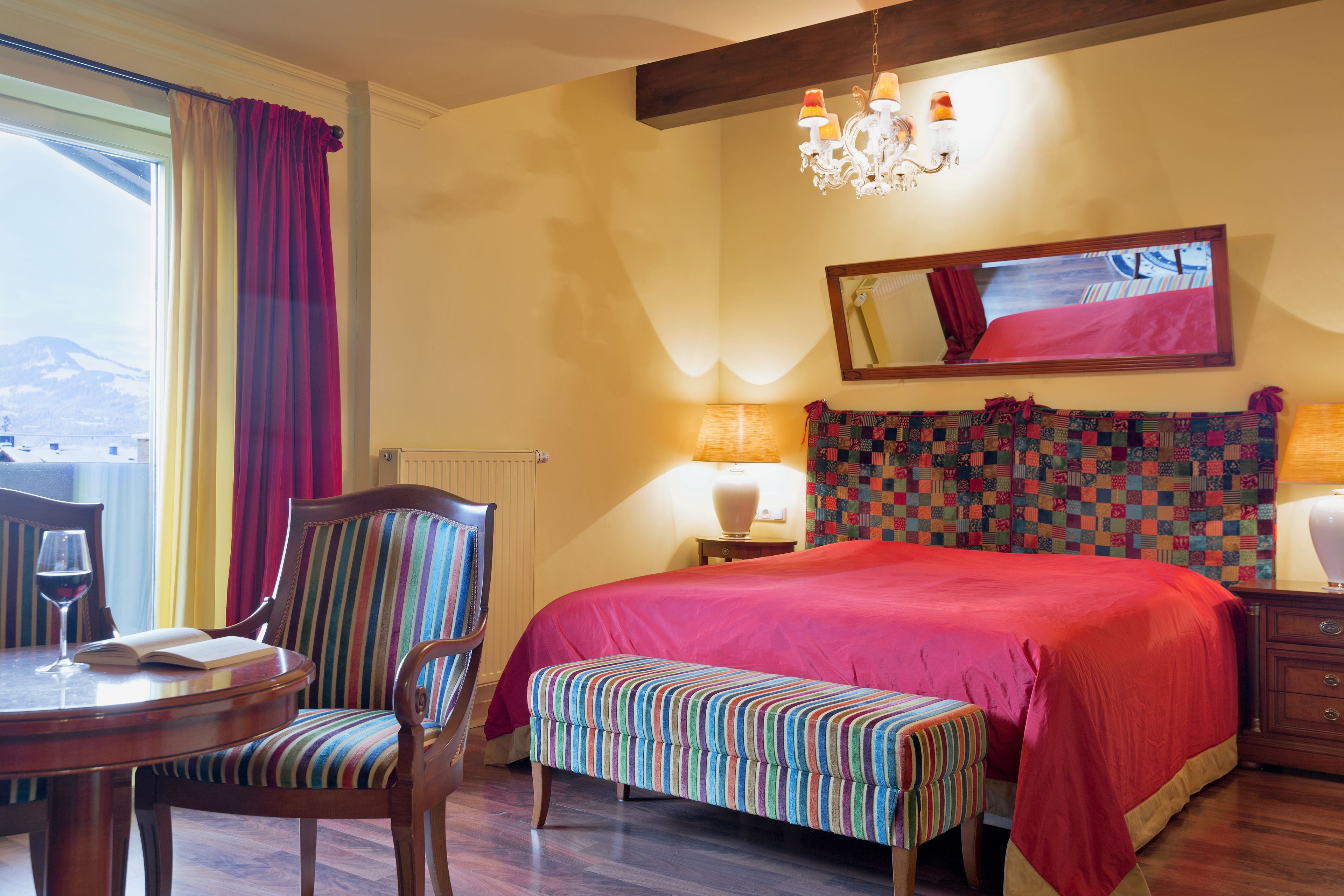 A hotel room with a large bed, a dining table with chairs, a bench, a chandelier, a mirror, and a view of the outdoors.