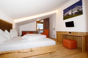 A modern bedroom with a wooden bed frame, a small orange ottoman, a flat-screen TV mounted on the wall, and a large window with curtains.