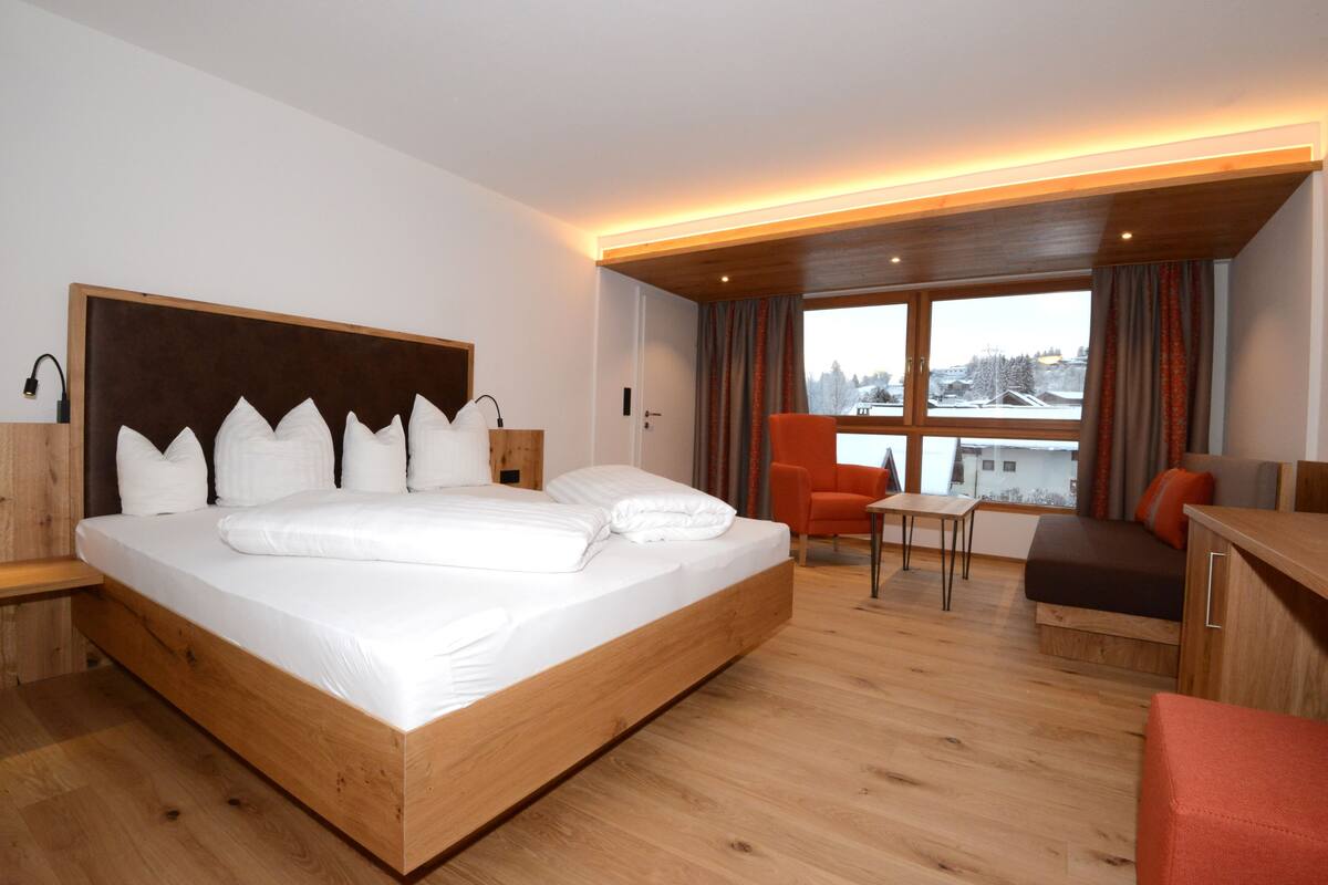 A modern hotel room with a large bed, a wooden headboard, a small table, a sofa, and a view of the city through large windows.
