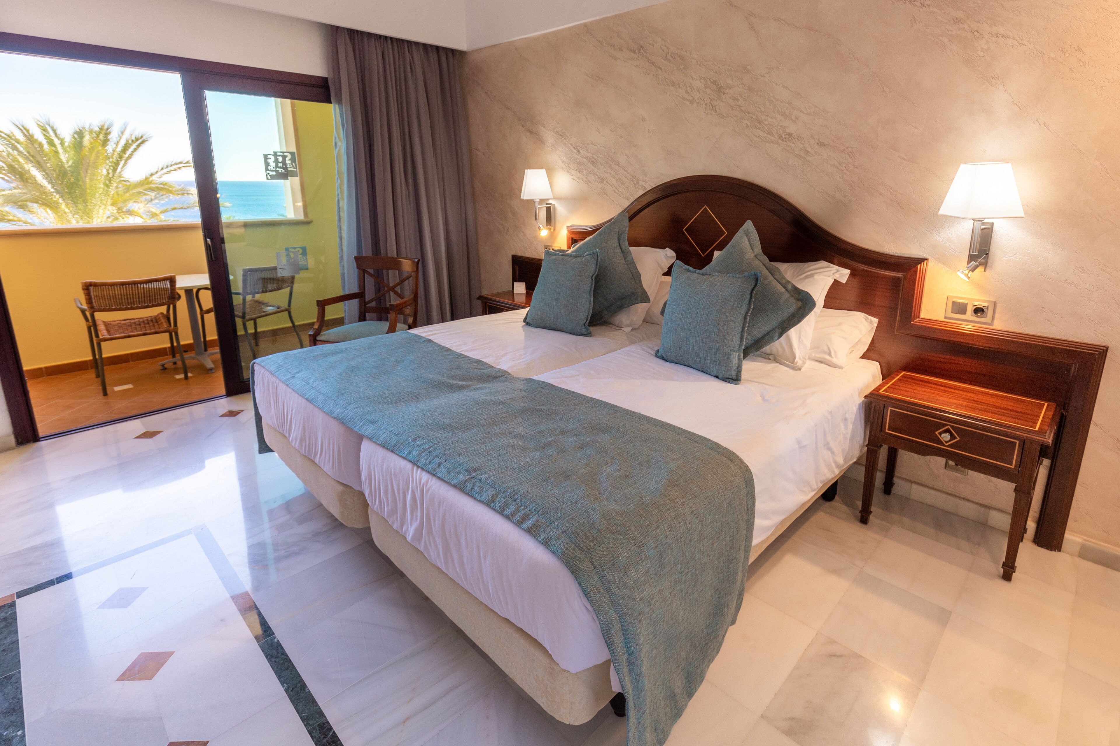 Habitación de hotel con una cama grande, dos mesitas de noche, un buró y un balcón con una silla, con vista al mar.
