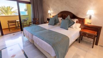 Habitación de hotel con una cama grande, dos mesitas de noche, un buró y un balcón con una silla, con vista al mar.