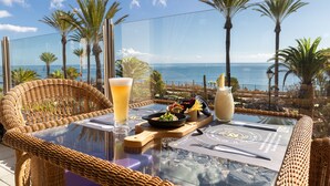 Uma mesa de jantar com tampo de vidro, rodeada por cadeiras de vime, com vista para a costa, palmeiras e o mar.