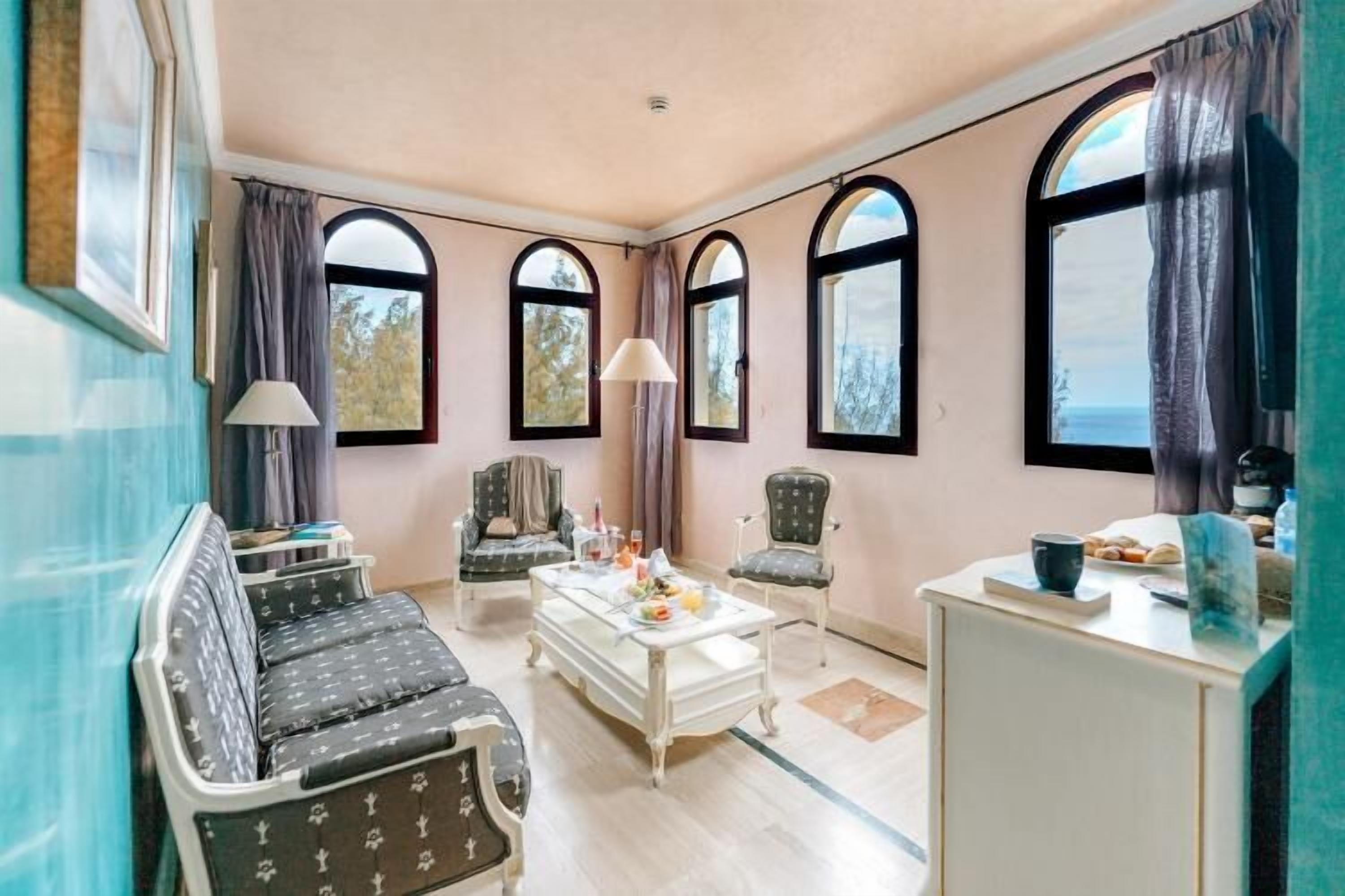 A room with a sofa, chairs, a small table, and three arched windows with curtains.