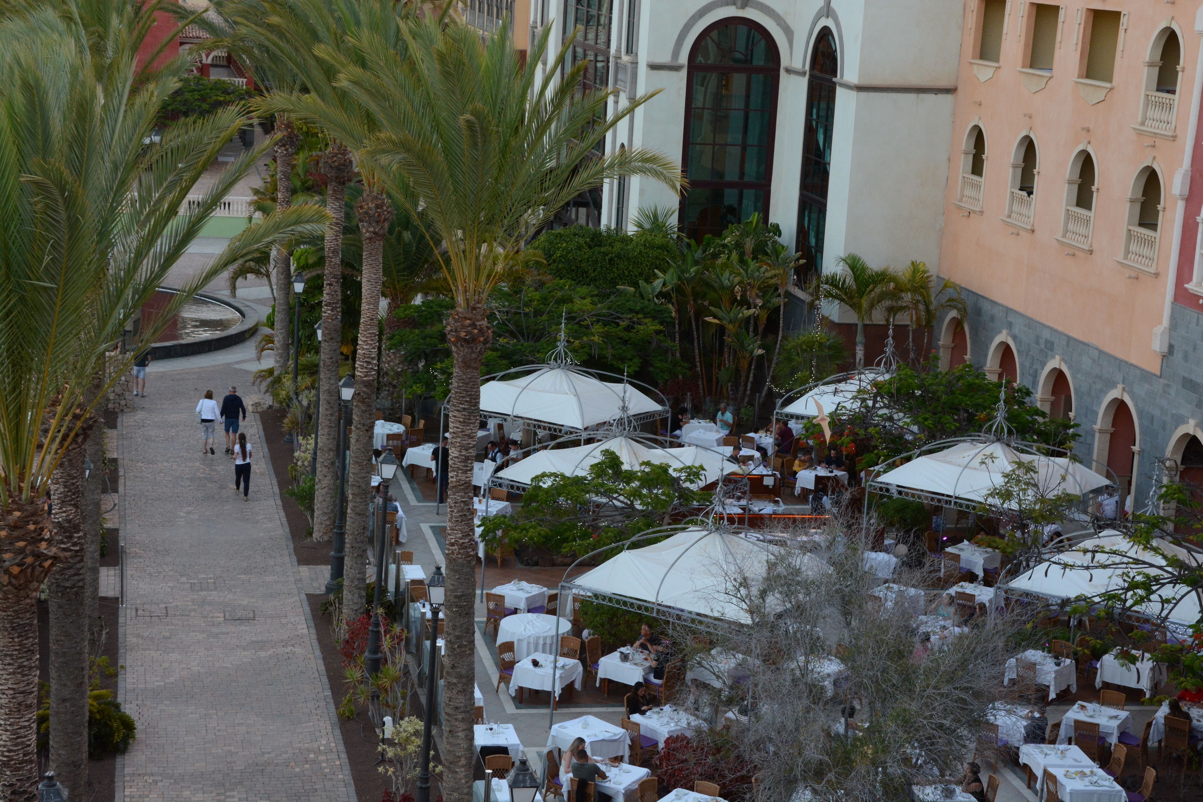 Un café al aire libre con mesas y sombrillas, rodeado de palmeras y un edificio con arcos.
