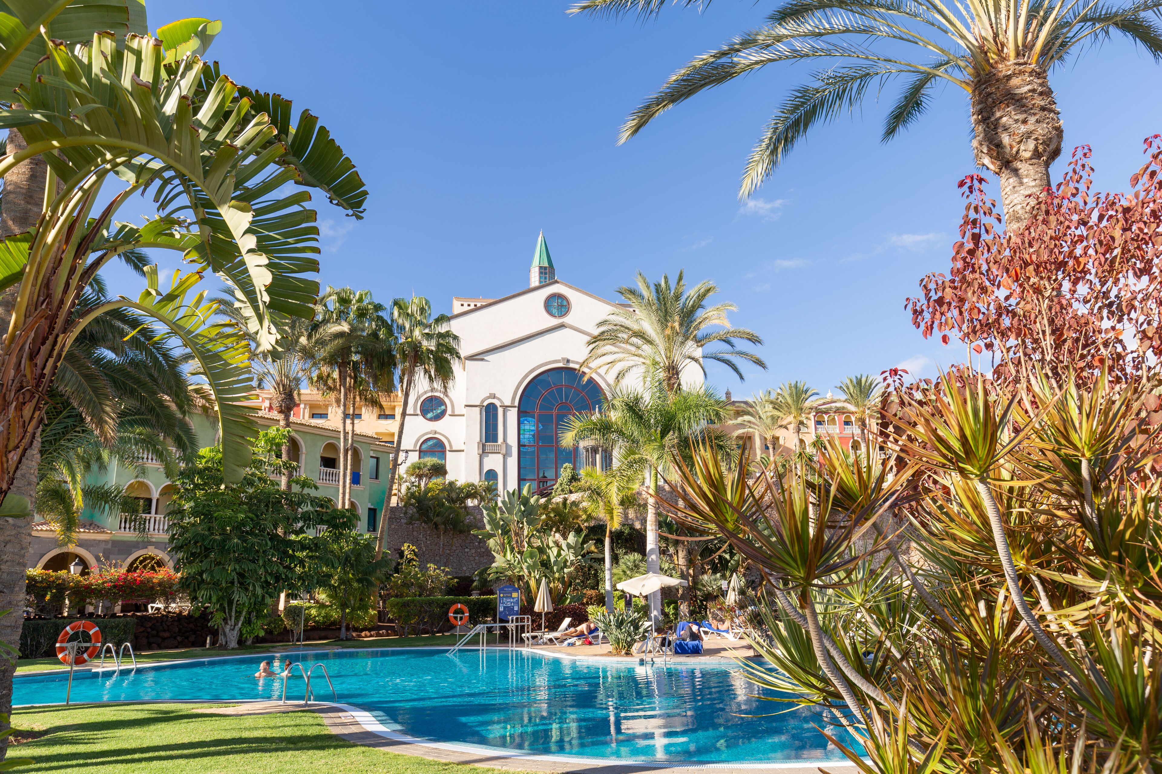 Un área de piscina con un edificio grande al fondo, rodeada de palmeras y otras plantas tropicales.
