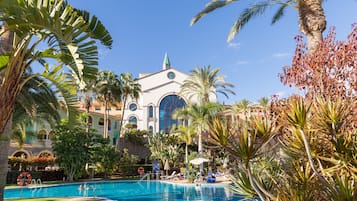 Un área de piscina con un edificio grande al fondo, rodeada de palmeras y otras plantas tropicales.