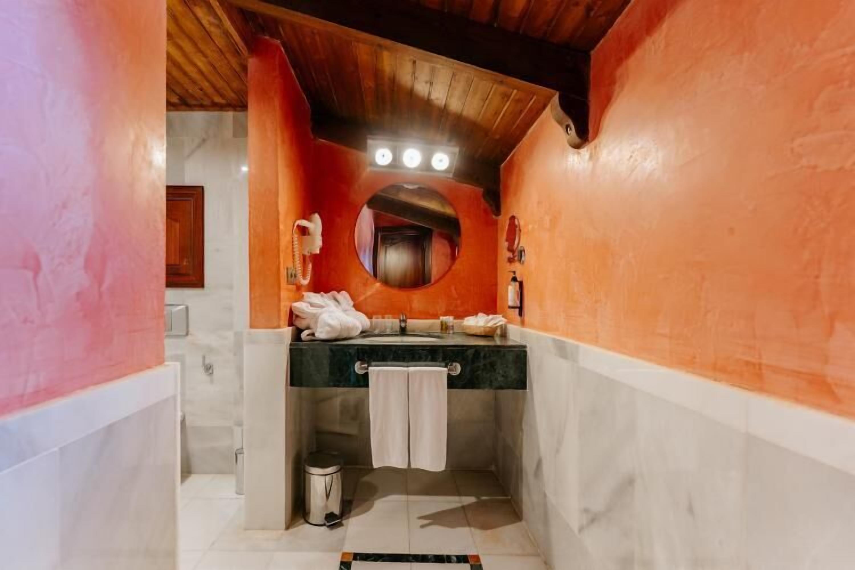 Ein Badezimmer mit Holzboden, einem runden Spiegel, einem Waschbecken mit Handtuch und einem Mülleimer.