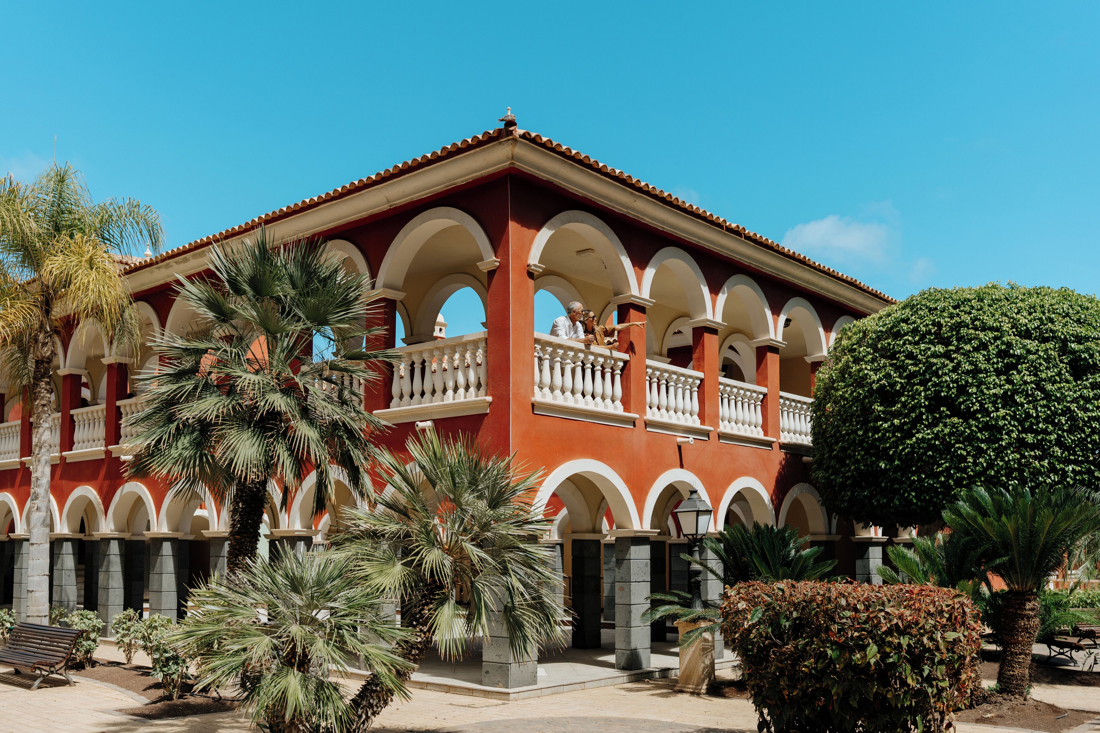 Un edificio rojo con arcos y balcones blancos, rodeado de palmeras y un jardín cuidado.