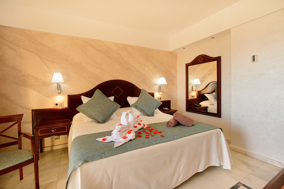 A hotel room with a neatly made bed, two nightstands with lamps, a wooden chair, and a large mirror on the wall.