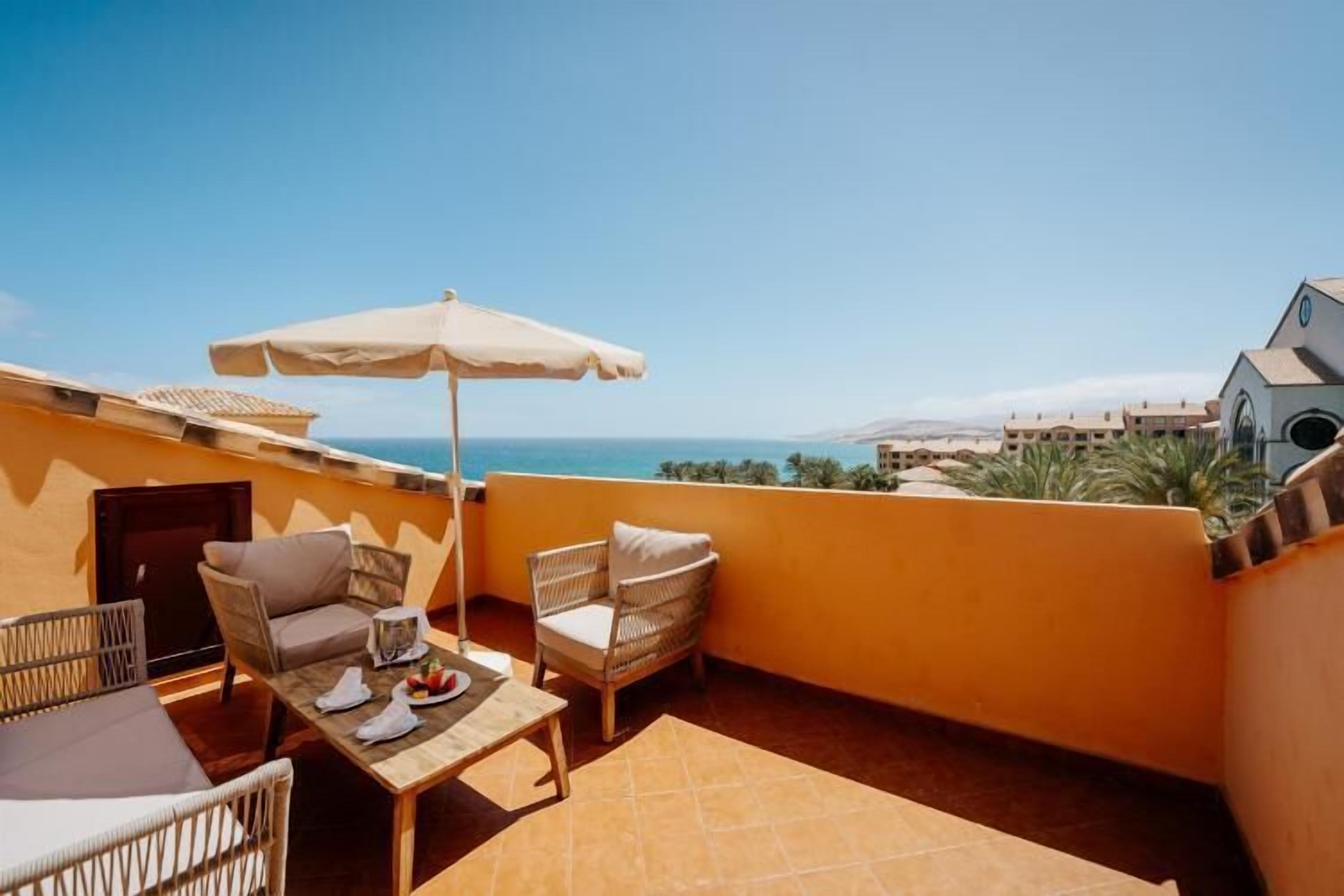 Un balcon avec une table, des chaises et un parasol, donnant sur une zone côtière avec des bâtiments.