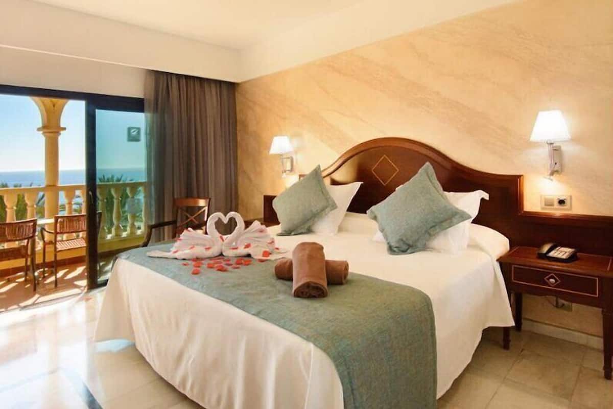 A hotel room with a bed, a nightstand, a lamp, a view of the ocean, and a balcony with chairs.