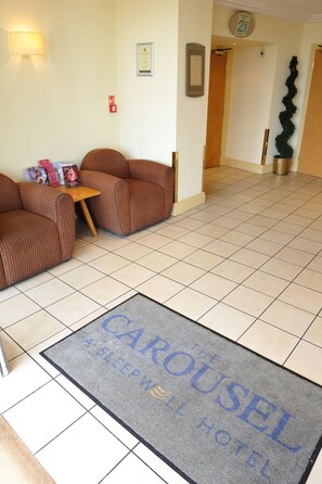A hotel lobby with a reception desk, two patterned armchairs, a doormat, and a potted plant. - Carousel Hotel (Blackpool)
