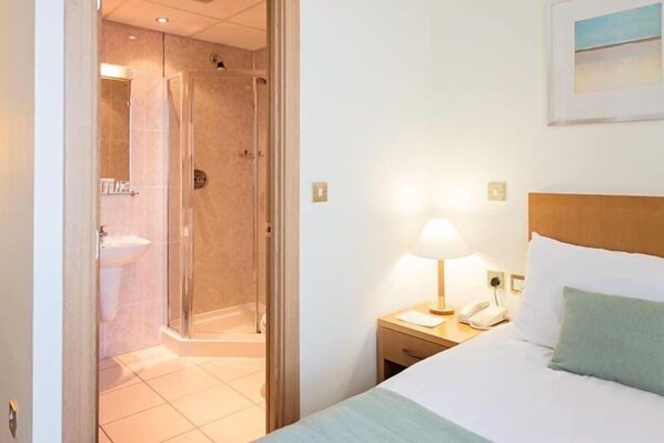 A hotel room with a bed, a nightstand with a lamp, a phone, and a shower area. - Carousel Hotel (Blackpool)