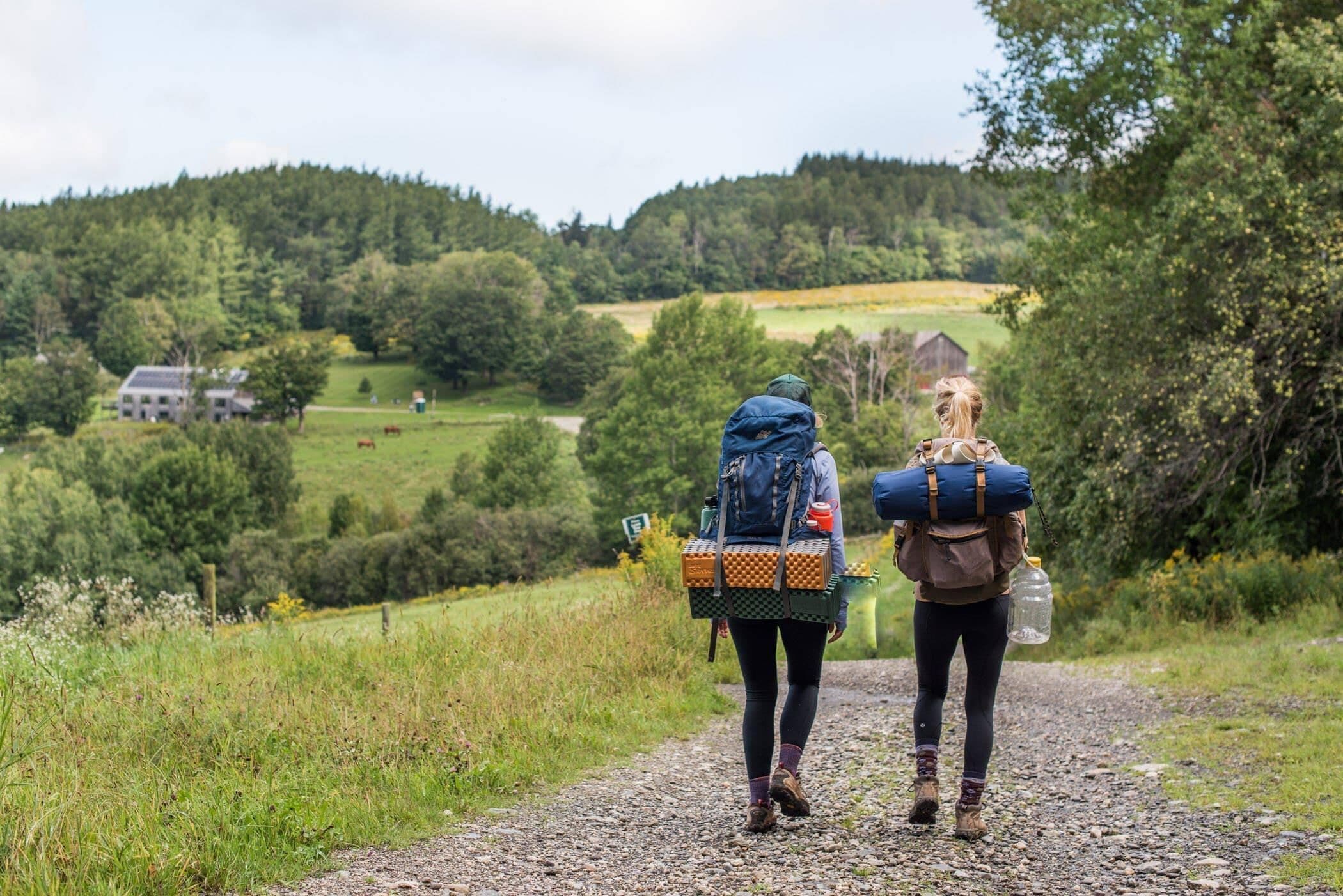 Zwei Personen gehen auf einem Kiesweg durch eine ländliche Gegend mit viel Grün und einem Haus im Hintergrund.