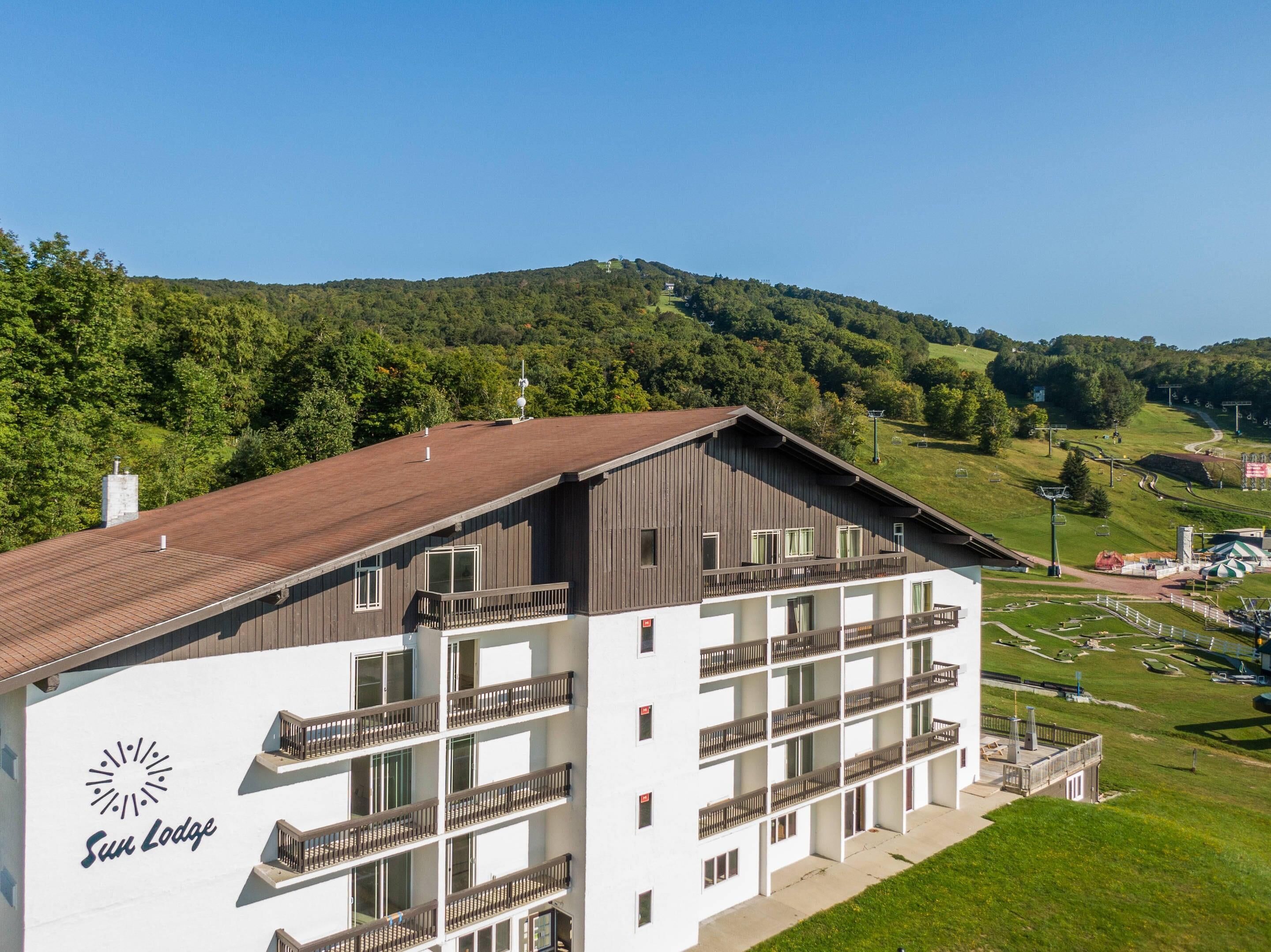 A multi-story hotel with balconies, a ski slope in the background, and a clear sky.