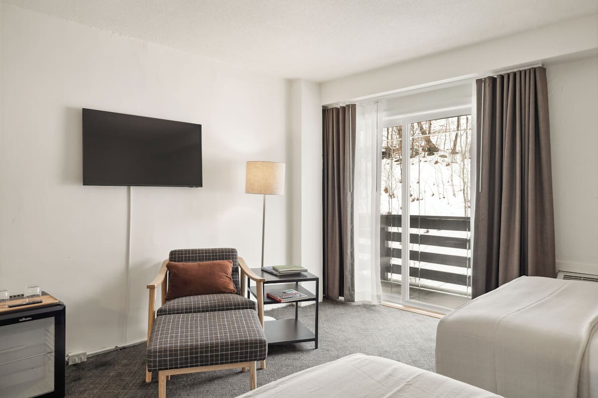 A hotel room with a bed, a chair, a small table with a lamp, a flat-screen TV mounted on the wall, and a window with curtains.