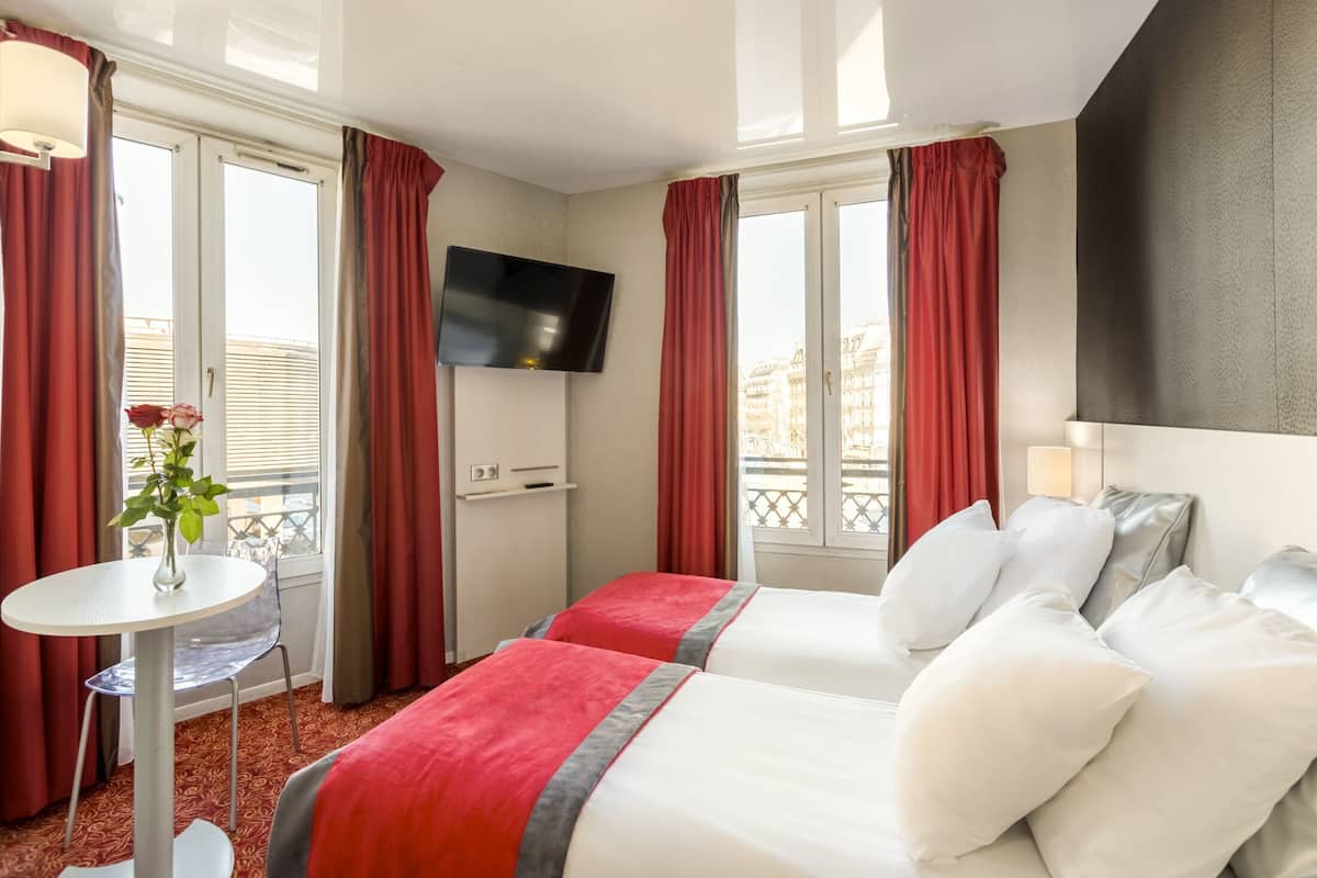 Habitación de hotel con dos camas, un televisor, una mesita con un florero y un balcón con cortinas rojas.