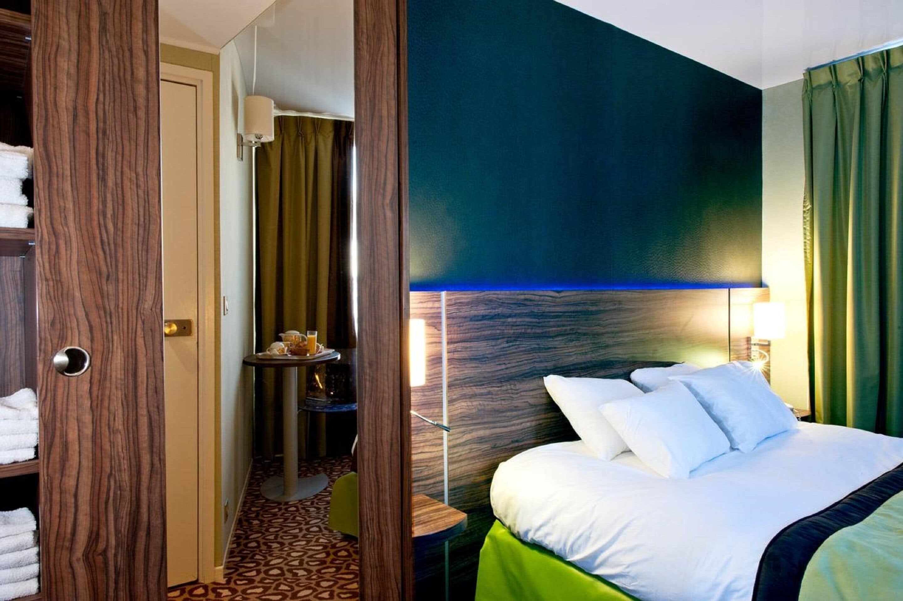 A hotel room with a bed, a small table with a lamp, a wall-mounted TV, and a door leading to a bathroom.