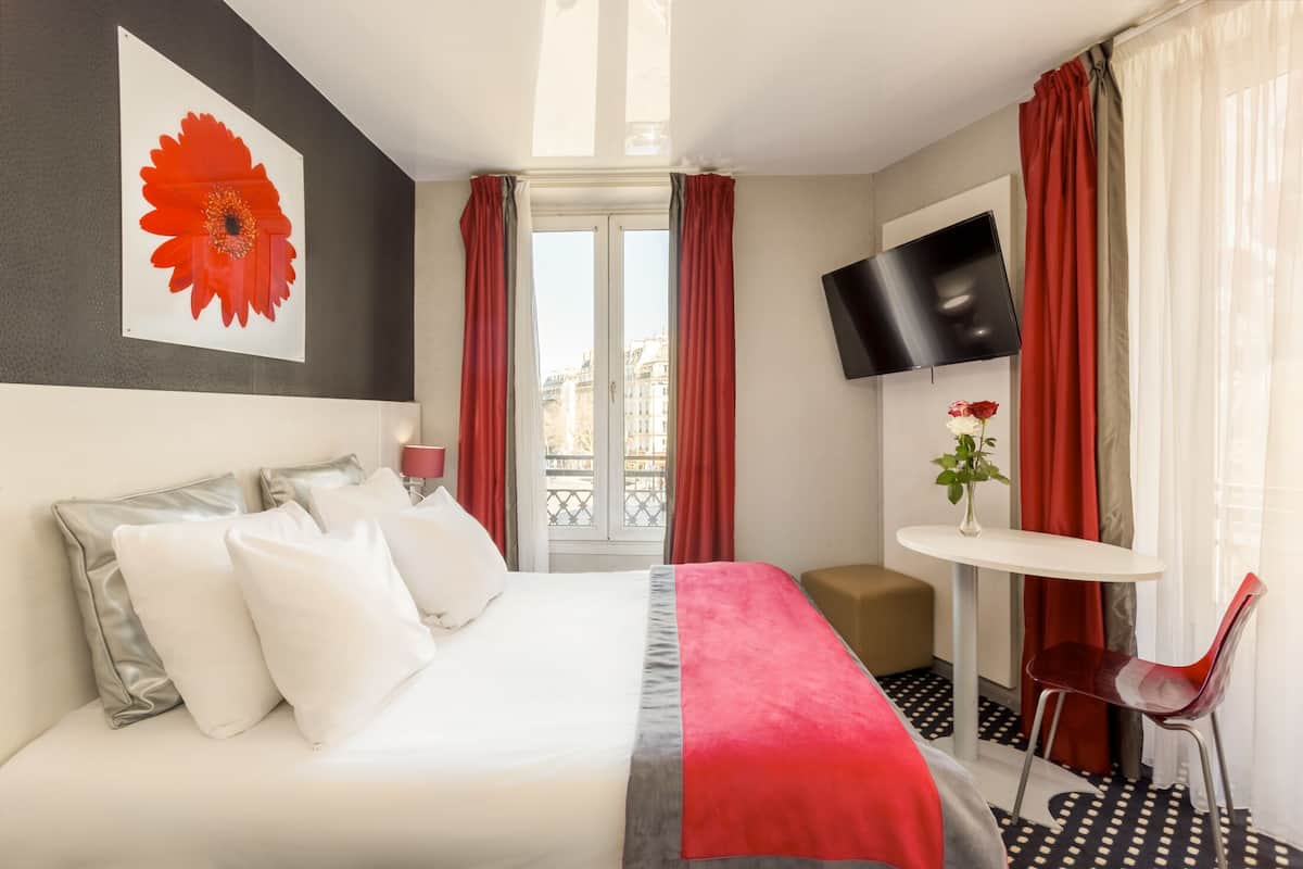 Una habitación de hotel con una cama, un televisor, una mesita con un florero y una silla.