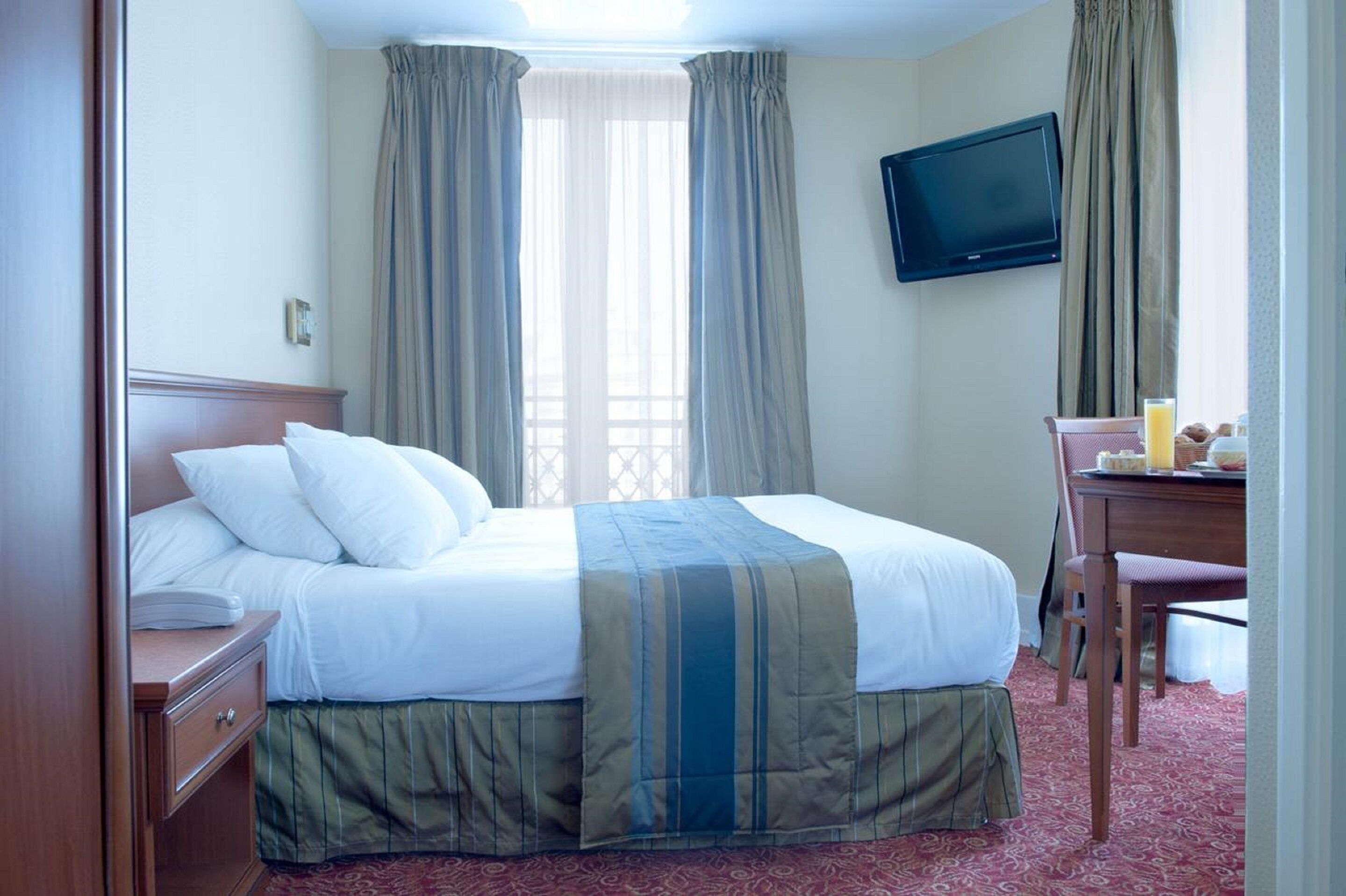 A hotel room with a bed, a desk with a chair, a television, and a window with curtains.