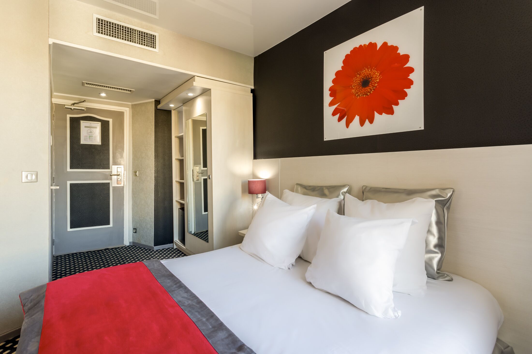 A hotel room with a bed, a red throw, a large flower artwork, and a bathroom visible through an open door.