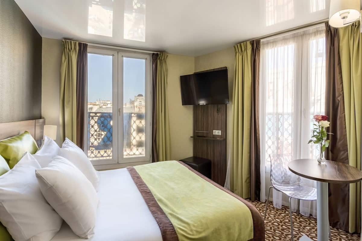 Habitación de hotel con cama, televisor, una mesita con un jarrón y vistas al balcón.