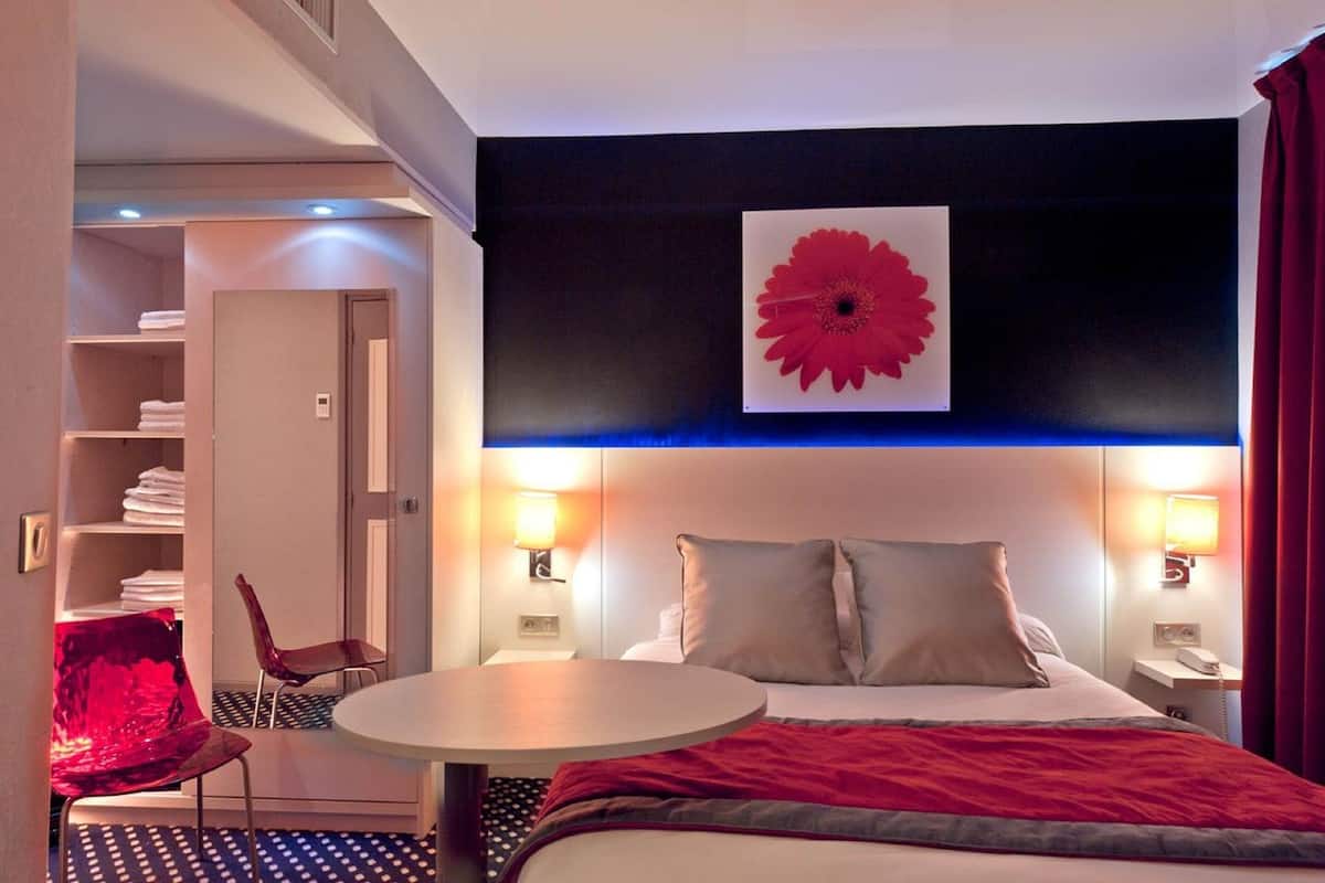 A hotel room with a bed, bedside tables, a round table, a chair, a wardrobe, and a large red flower decoration on the wall.