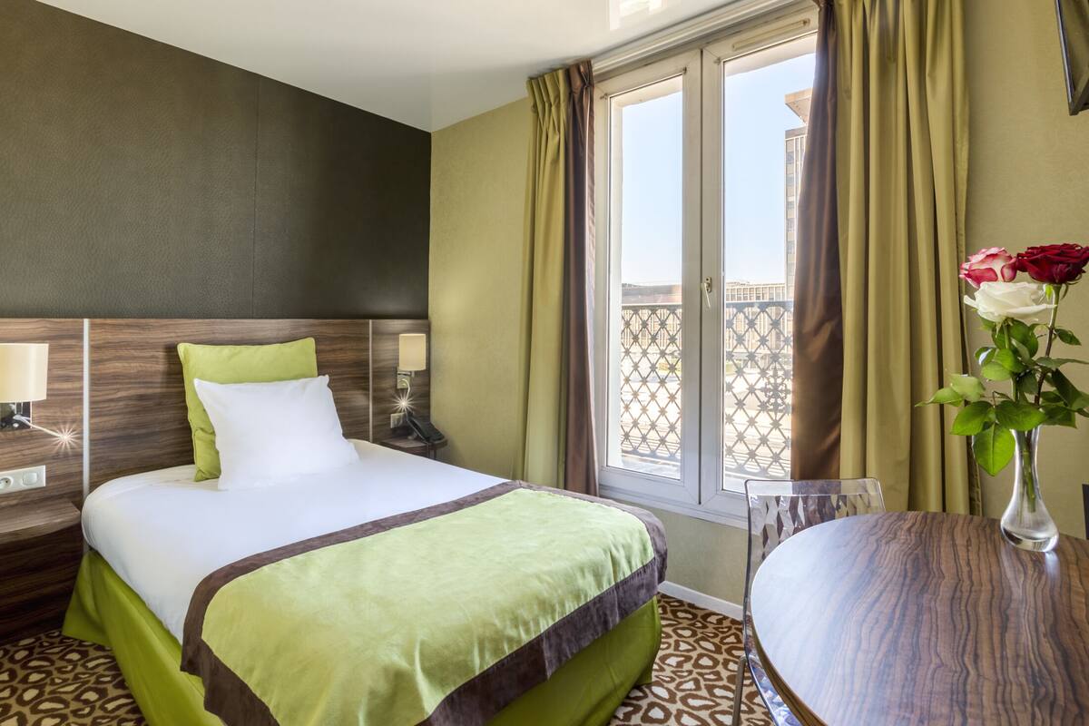 Habitación de hotel con cama, una mesa, un florero con flores y vista al exterior.