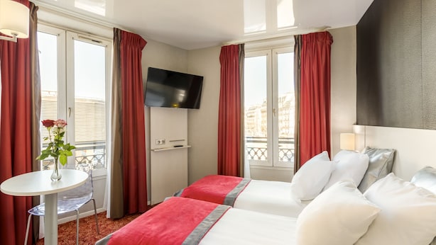 A hotel room with two beds, a TV, a small table with a vase of flowers, and large windows with red curtains. - Hotel Berkeley (Paris)