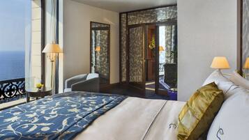 A hotel room with a large bed, a chair, a small table with a lamp, and a view of the sea.