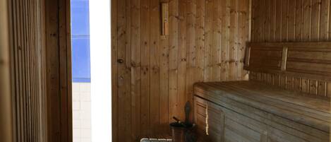 Interior de una cabaña de madera con un banco, una mesita con una olla y una puerta que da al exterior.
