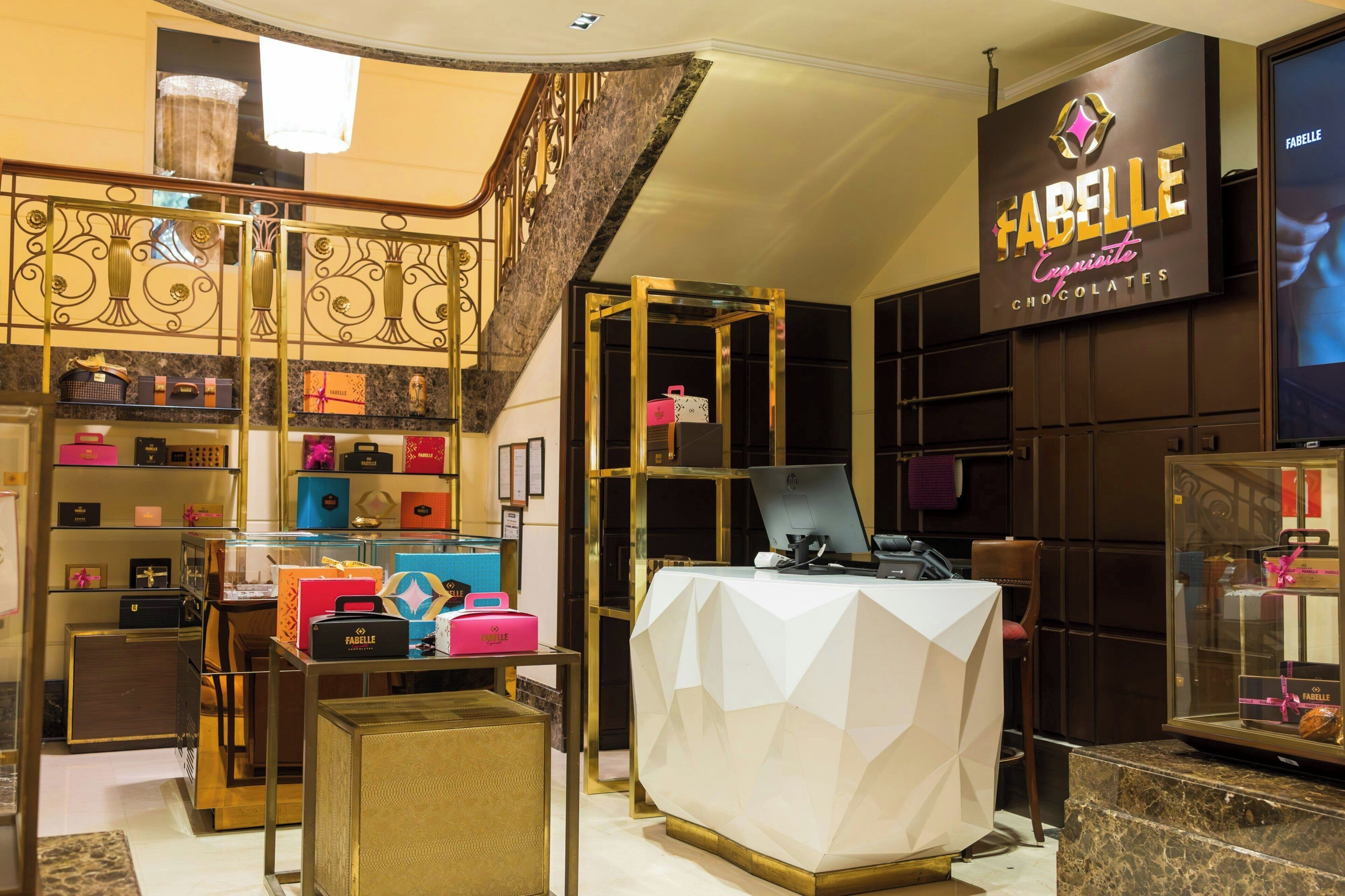 A luxury chocolate store interior with a geometric-patterned reception desk, shelves of chocolate boxes, and a staircase with a decorative railing.