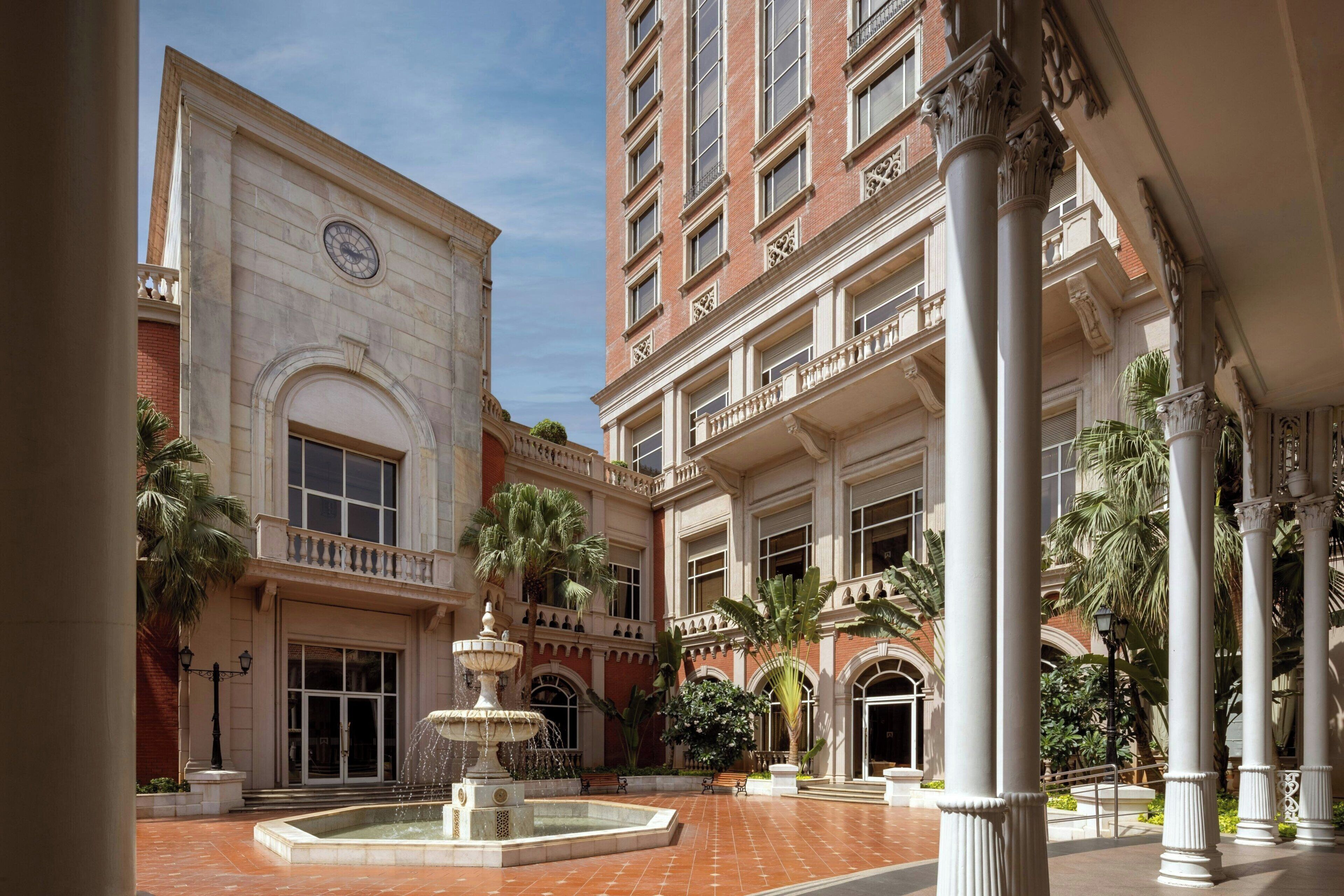 A courtyard with a fountain, palm trees, and classical architecture.