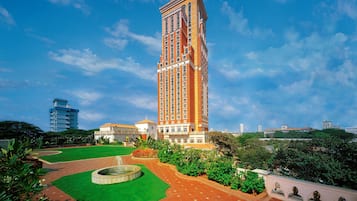 Un edificio alto de ladrillo rojo con una torre de reloj, rodeado de un jardín ajardinado con una fuente, bajo un cielo azul despejado.