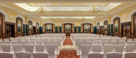 Un salón amplio con filas de sillas blancas y una alfombra roja, que presenta candelabros y puertas decoradas.