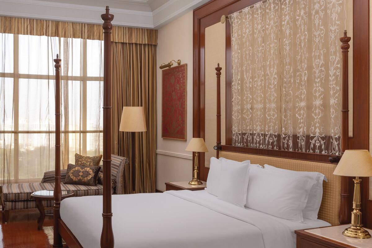 A four-poster bed with white linens, two bedside tables with lamps, a large window with curtains, and a patterned rug.