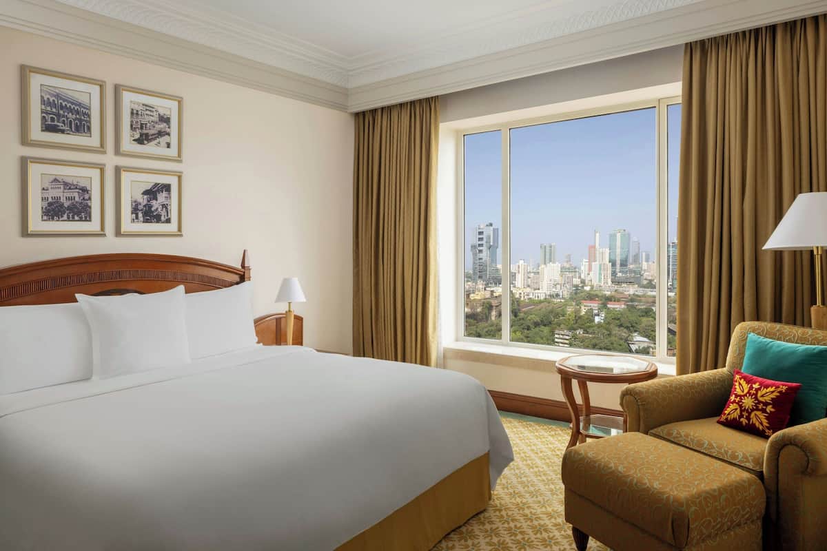 A hotel room with a large bed, a chair, a small table, and a window with a city view.