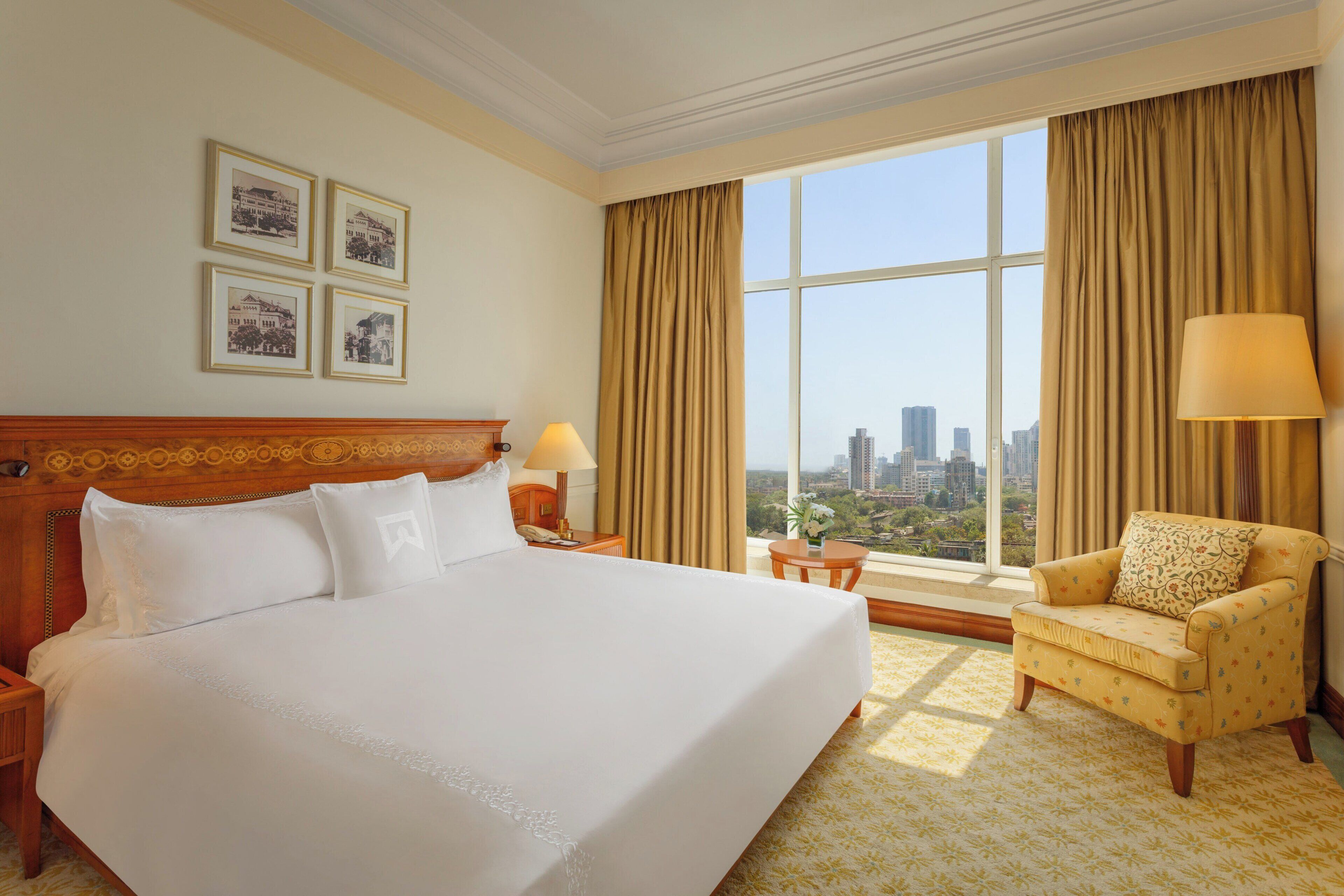 A hotel room with a large bed, a chair, a nightstand, a lamp, and a window with a city view.
