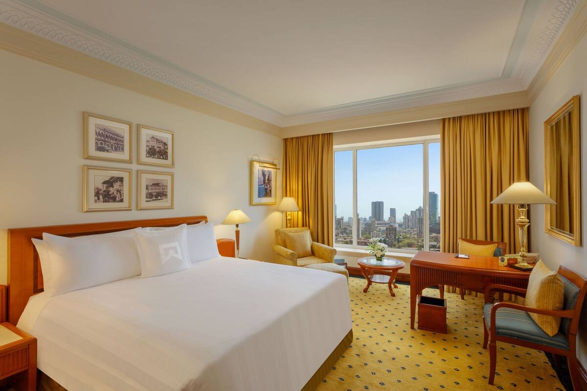 A hotel room with a large bed, a desk, two chairs, a small table, and a large window with a city view.