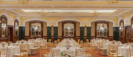 Uma sala de banquetes com mesas redondas, cadeiras douradas, lustres e colunas de mármore.