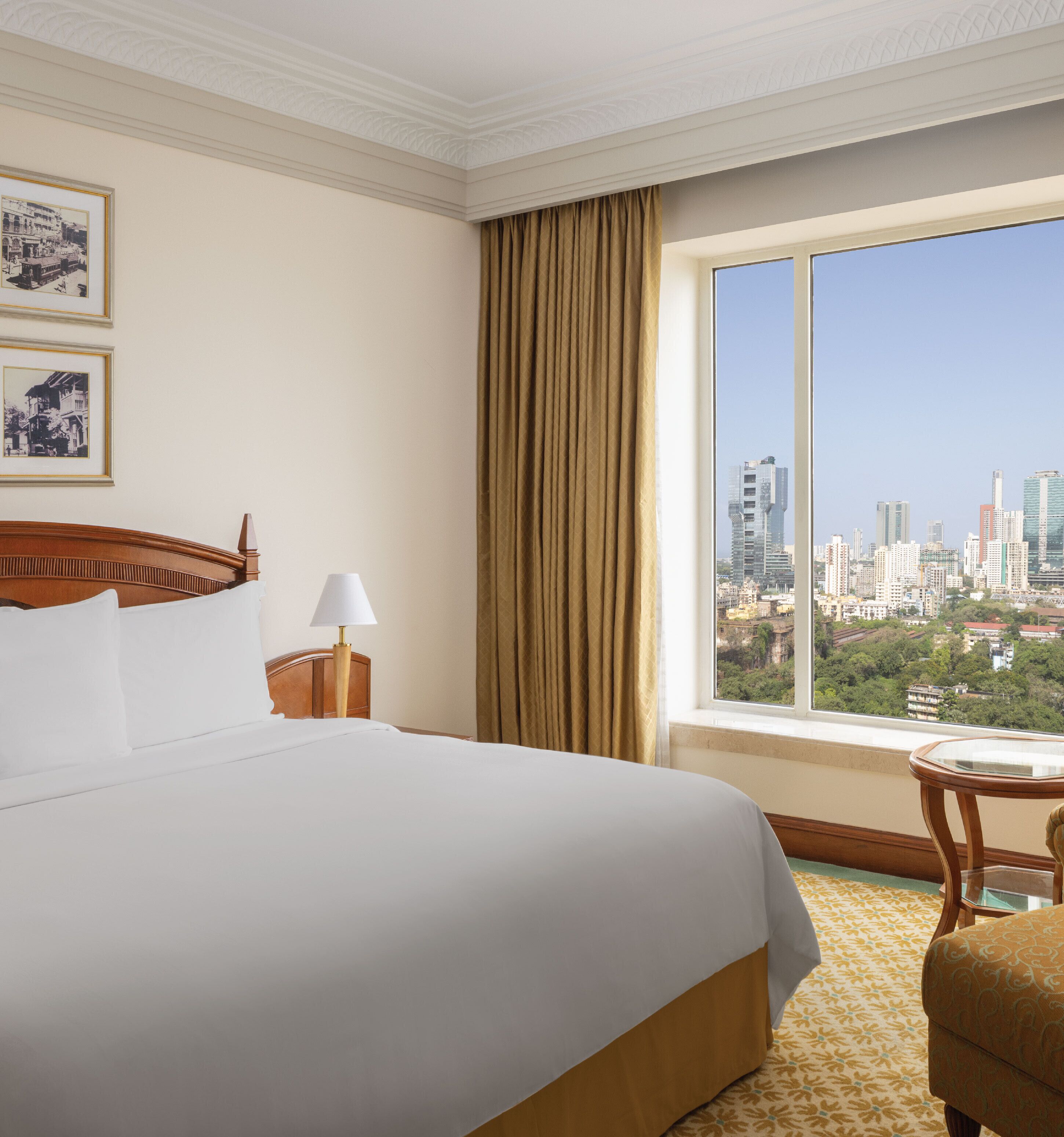 Habitación de hotel con una cama grande, una mesita de noche con lámpara, una silla y una ventana con vista a la ciudad.