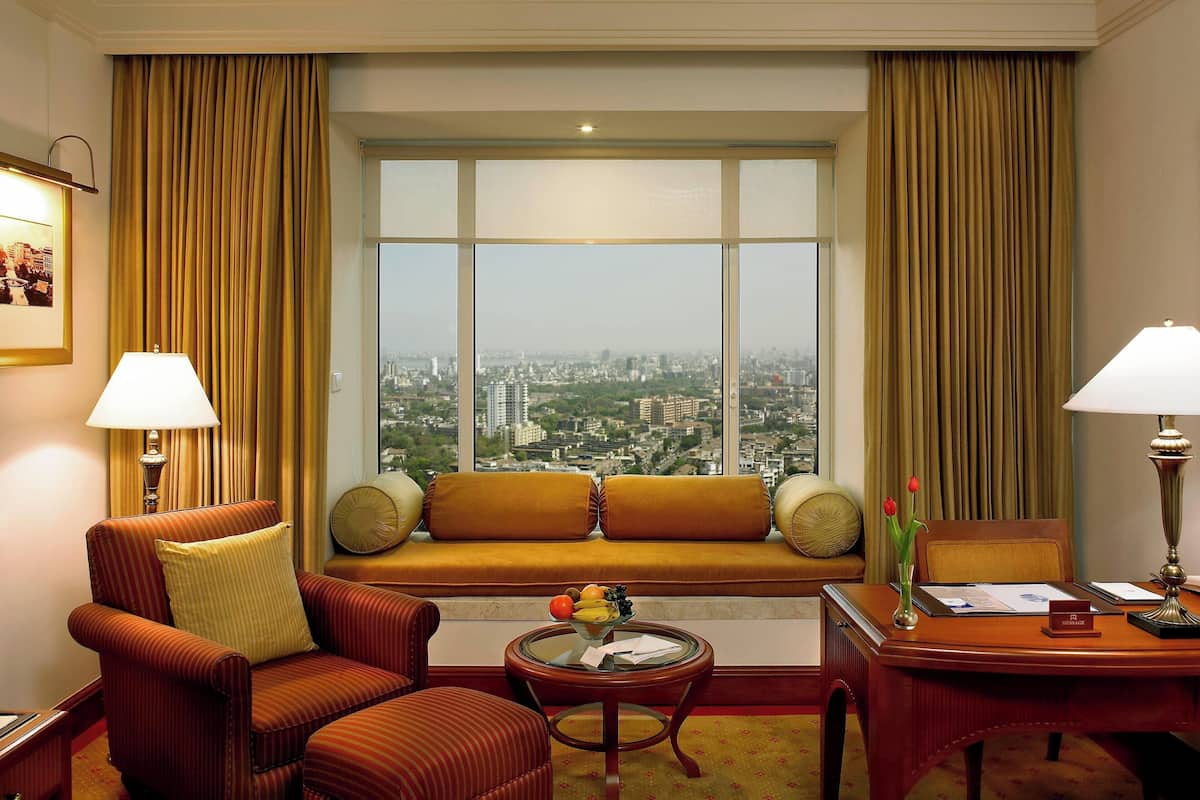 A hotel room with a large window, a sofa, a chair, a desk, and a lamp.