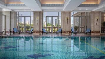 An indoor swimming pool with clear glass walls, blue lounge chairs, and a city view.
